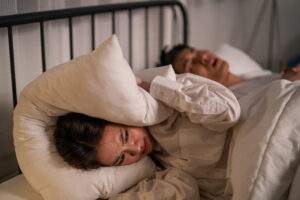 Asian woman feel frustrated from husband snoring while sleep at night. Attractive insomnia angry wife lying down on bed, cannot sleep due to noise and put cozy blanket on her ears in bedroom at home.